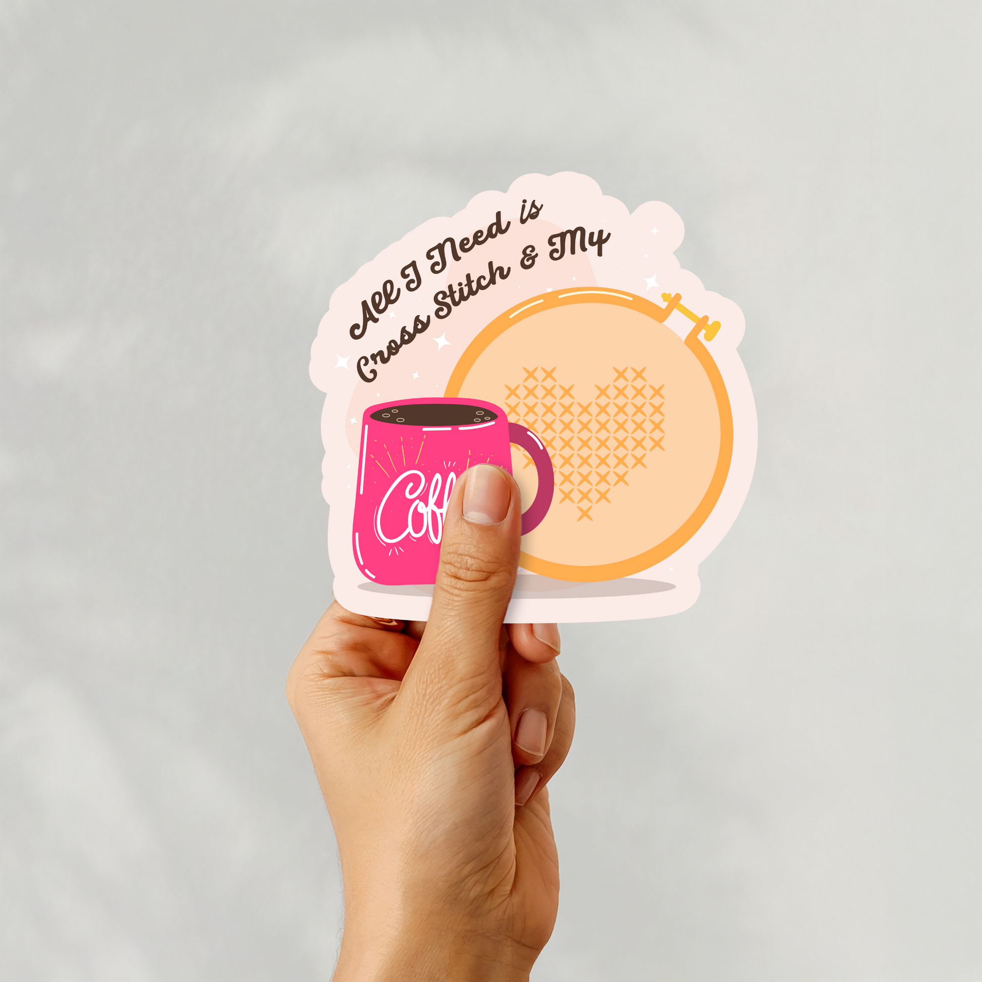 Hand holding a “All I Need is Cross Stitch & My Coffee” sticker with heart cross-stitch hoop and pink coffee mug illustration