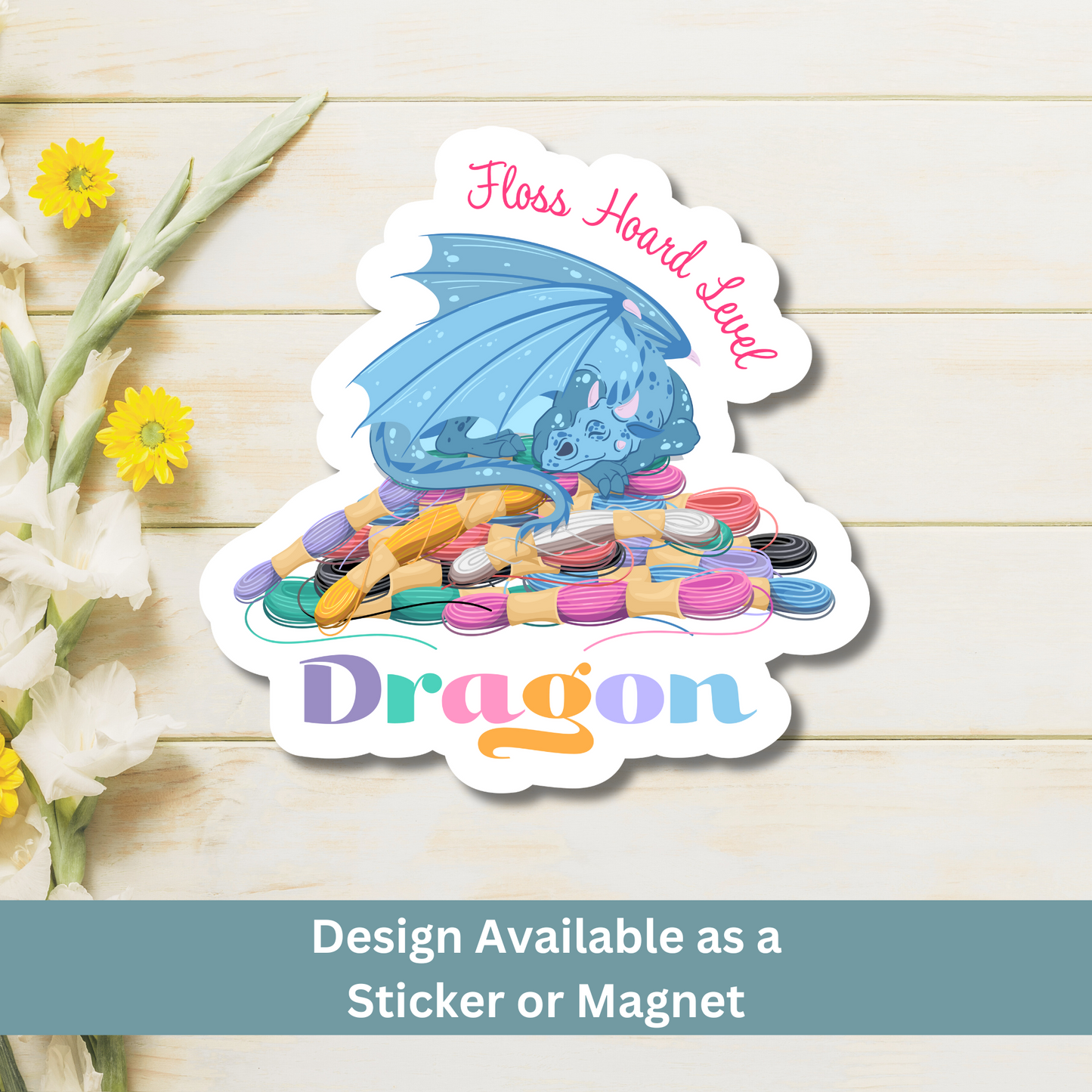 A magnet of a dragon sitting on embroidery floss on a wooden surface, text says "Floss Hoard Level Dragon"