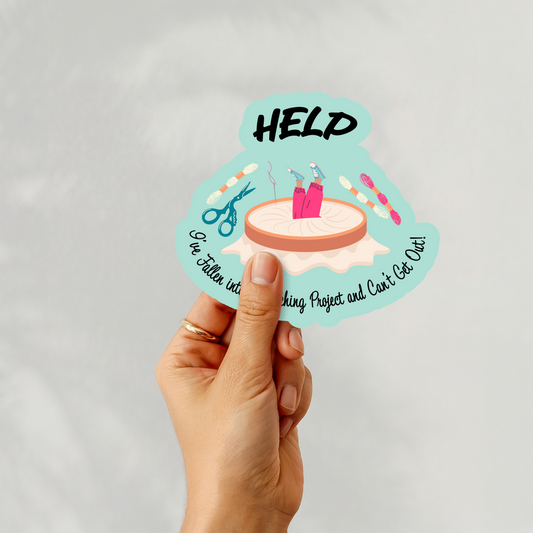 Hand holding a humorous sticker with “Help I’ve Fallen Into a Stitching Project” and cross-stitch tools illustration