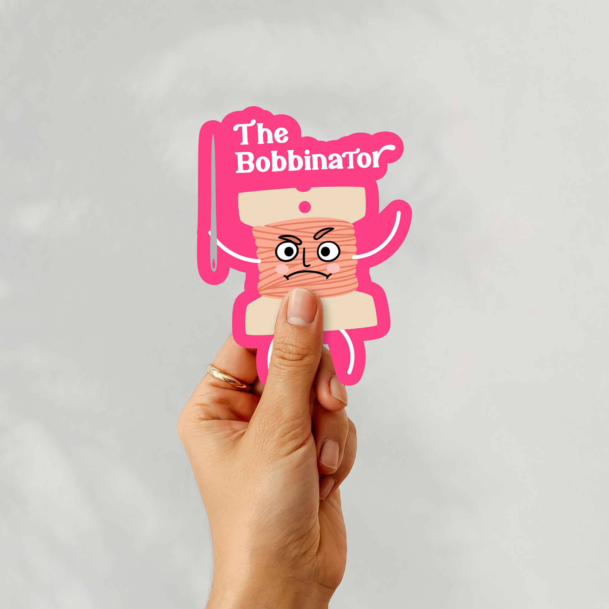 Hand holding ‘The Bobbinator’ sticker featuring cartoon bobbin character with needle