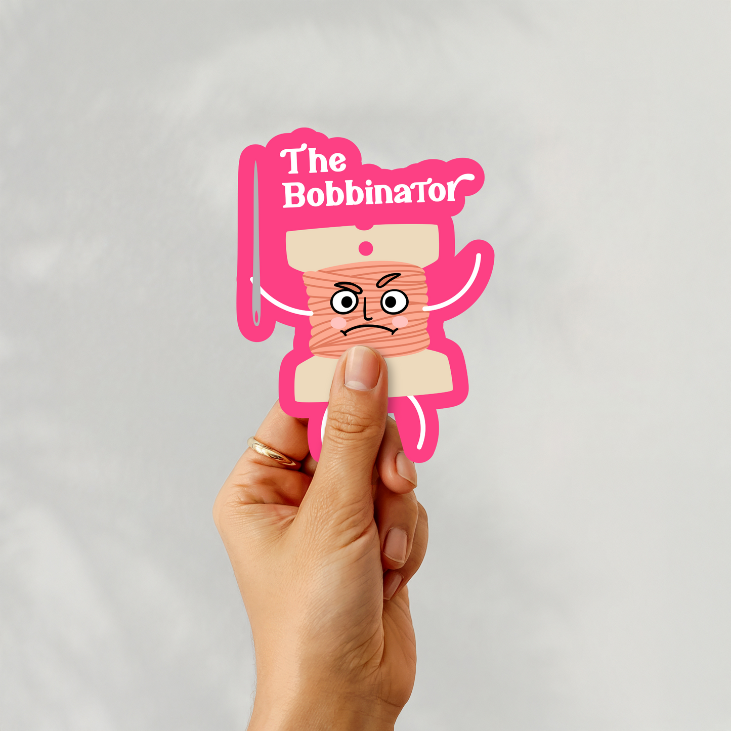 Hand holding ‘The Bobbinator’ sticker featuring cartoon bobbin character with needle