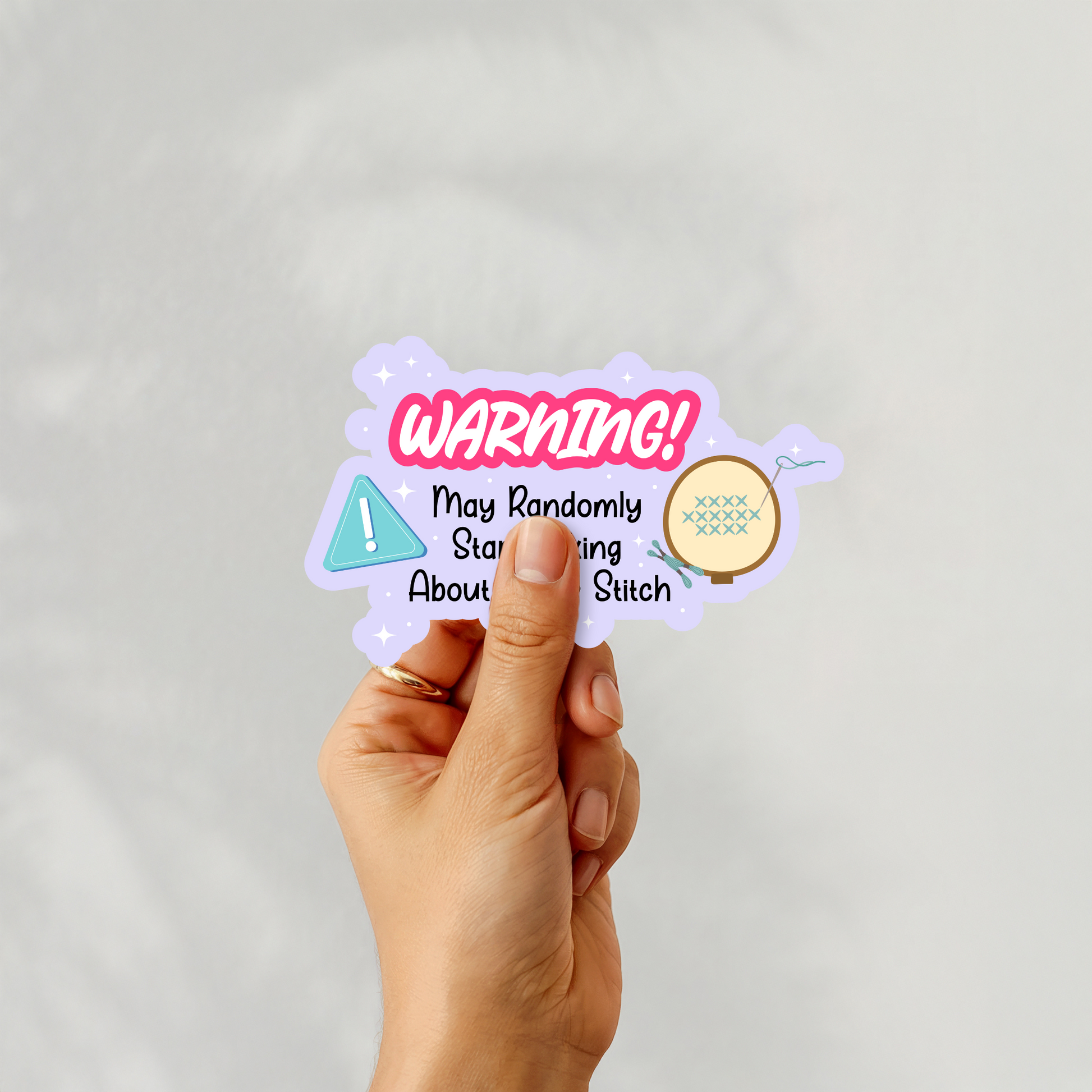 A hand holding a warning-themed sticker with the phrase “May Randomly Start Talking About Cross Stitch”